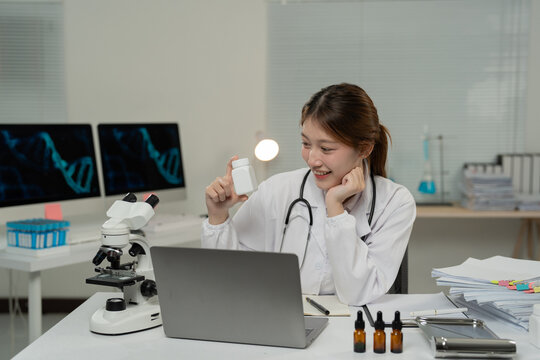 Asian female doctor in lab coat and stethoscope shows a medicine bottle during a video conference in her laboratory, with microscope and dna on screens