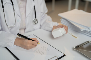 Doctor is writing medical prescription holding a pill bottle while sitting at a desk in hospital, healthcare and medical concept