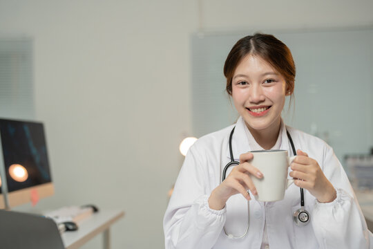 Young female doctor wearing lab coat and stethoscope, enjoying a coffee break in her modern office, showcasing a positive and healthy work-life balance