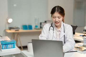 Young female doctor wearing lab coat and stethoscope working on laptop in her bright modern office or laboratory, smiling and looking at the screen