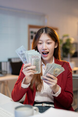 Asian businesswoman in red jacket is happily counting us dollar banknotes at her office desk, celebrating financial success, investment profit, or bonus