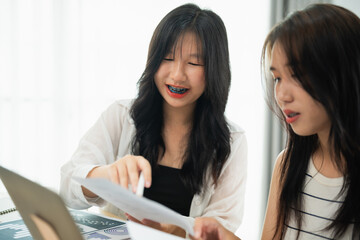 Two Asian women are sitting at a table looking at a laptop. One of them is pointing at something on the screen