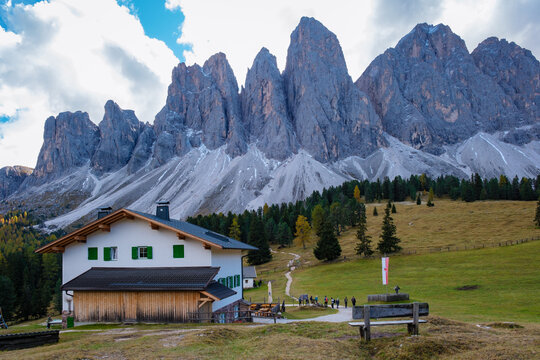 Under the vast sky, a quaint chalet sits in the foothills of the majestic Dolomites. Hikers wander along a winding trail, surrounded by dramatic mountain scenery and vibrant autumn colors.