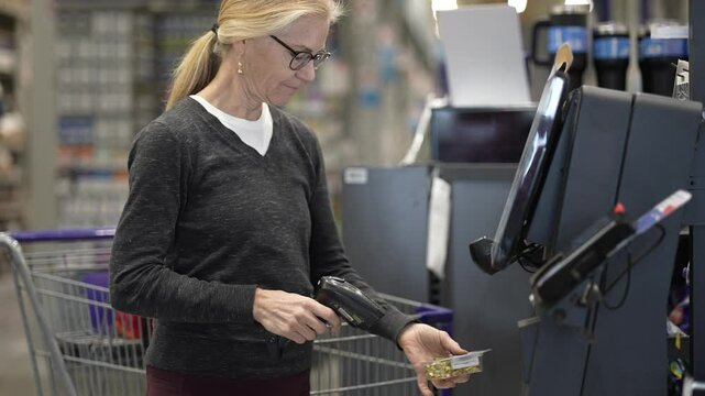 A woman with blonde hair and glasses scans items at a selfcheckout station in a retail store. She holds a barcode scanner and is looking down at the item she is scanning.