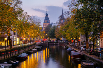 Fototapeta premium Golden leaves flutter above tranquil canals, reflecting the warm lights of bridges and buildings as twilight envelops Amsterdam.