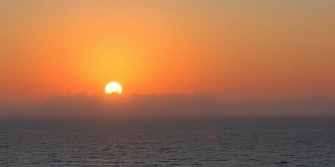 The sun rises above the calm and tranquil sea with a thin layer of fog drifting across its surface, soothing scenery, peaceful atmosphere