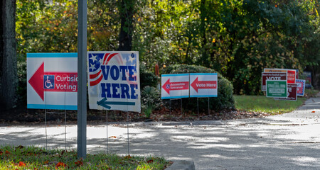 Vote Here signs directing voters to polling places, with secondary signs and candidate signs in soft focus in background.