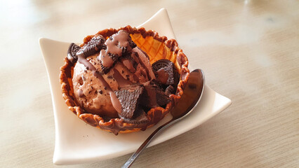 Chocolate ice cream with chocolate sauce, decorated with biscuit crumbs on top, served in a waffle bowl.