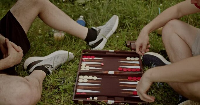 Two friends enjoy a game of backgammon in the park on a beautiful sunny day. They relax and strategize on the grass.