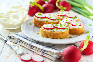 Appetizing crunchy baguette slices with cream cheese and radish slices and freshly chopped chives on a white wooden table.