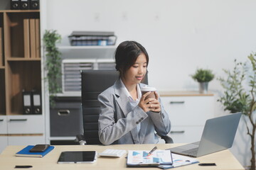 Obraz premium Successful Asian businesswoman smiling using laptop computer and holding coffee cup at office. Confident Asia businesswoman sitting happily in the office.