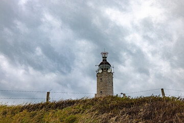 The historic lighthouse sits atop a grassy hill under a dramatic sky, guiding navigation along...