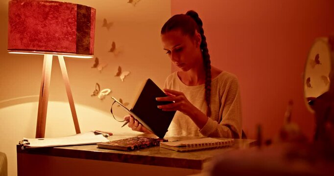 Young student works on school assignment late night in cozy dorm room. Girl prepares for exams, does homework. Education, learning, and studying concept.