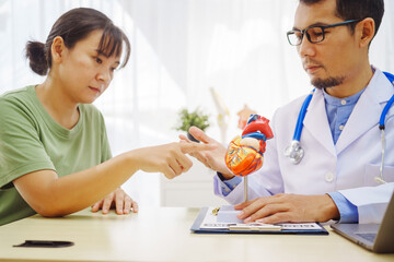 A doctor man sits at a desk in a hospital, explaining heart disease symptoms to a female patient. They discuss chest pain, palpitations, fatigue, dizziness, and the risks of myocardial ischemia