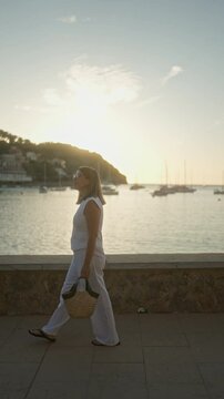 Woman walking along port de soller promenade at sunset with boats in the background, mallorca, spain, depicting serene travel and leisure vacation vibes with a coastal view