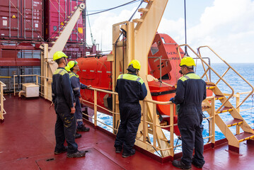 A group of merchant ship crew members stands together during an abandon ship drill, discussing the...