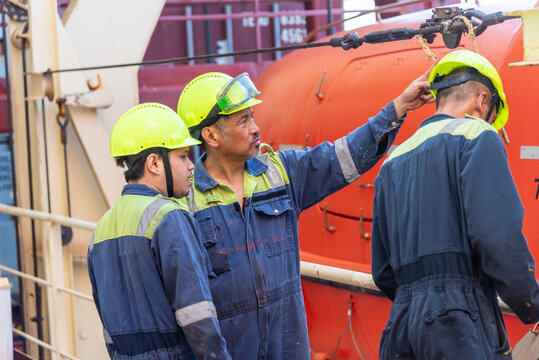 A group of merchant ship crew members stands together during an abandon ship drill, discussing the procedure for lowering the lifeboat. Their coordinated focus highlights the importance of teamwork.