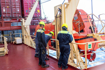 A group of merchant ship crew members stands together during an abandon ship drill, discussing the...