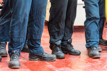 A close-up of the legs of a group of people, all dressed in work clothing and safety boots,...
