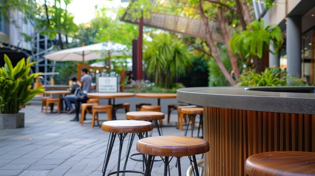 Wooden stools stand beside a bar in a modern outdoor cafe seating area in an urban green space, with customers enjoying the atmosphere in the background