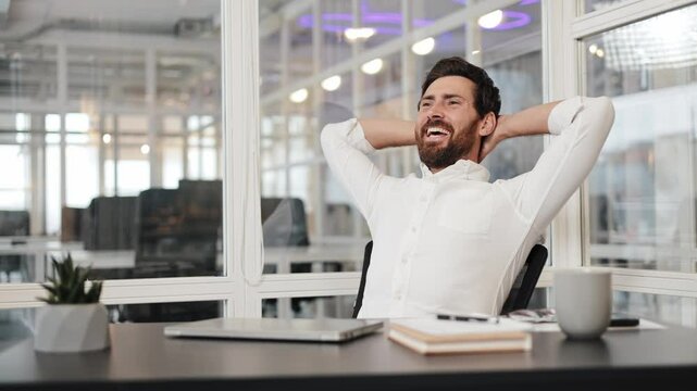 Satisfied businessman finishes working on his laptop, closes it, leans back, puts his hands behind his head, and laughs with joy and relief.