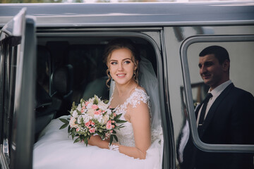 A bride is getting into a limousine with a man behind her. She is holding a bouquet of flowers