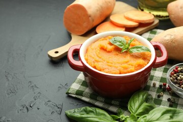 Tasty mashed sweet potato with basil in pot, fresh vegetables and spices on dark textured table, closeup