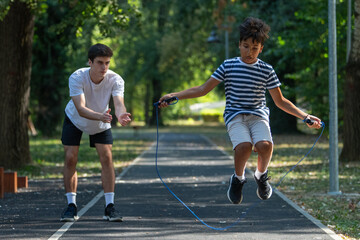 Young boy jumps rope outdoors with guidance from a trainer, emphasizing coordination and fitness. The photo highlights an energetic, fun training session in a natural, open setting.