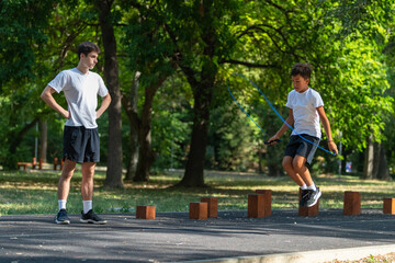 Boy practices jumping rope outdoors with a trainer, promoting fitness and coordination. This...