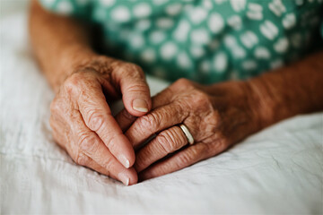 The hands of an elderly person rest gently on a soft surface, displaying intricate details of age, wearing a simple ring and suggesting a moment of reflection or peace indoors.