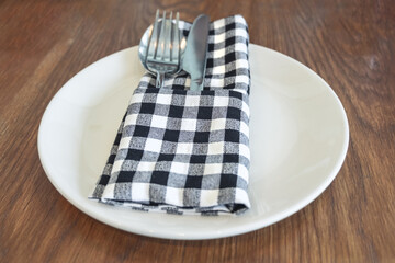 A neatly arranged table setting featuring a checkered napkin, fork, knife, and a simple white plate on a wooden surface.