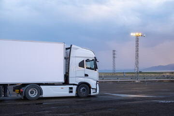 A big white truck parked at a service station at dusk to fill the fuel tank. The asphalt ground is wet from the rain. Concept of fossil fuels in the transportation of goods by road © LaMorenita