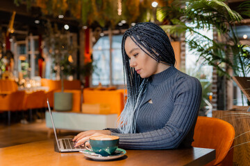 young woman with Afro braids is sitting in a coffee shop and working on a laptop