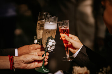  Three people raise decorated champagne flutes in a celebratory toast, with sparkling drinks, floral accents, and engraved details on the glasses.