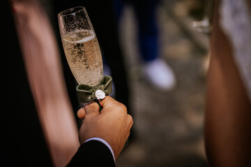 Close-up of a hand holding a champagne flute decorated with a small green bow and white flower, likely during a wedding or celebration toast.