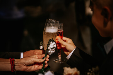  Three people raise decorated champagne flutes in a celebratory toast, with sparkling drinks, floral accents, and engraved details on the glasses.