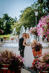 Fototapeta premium Bride and groom pose together, surrounded by vibrant flowers and fountains in an outdoor setting on a bright, sunny day..