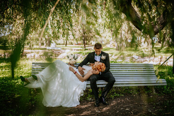  Bride rests on groom’s lap on a park bench, holding her bouquet, under a canopy of leafy...