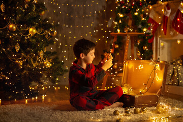 Child with shining Christmas Ornaments. Little Boy in Pajamas opening Vintage Suitcase full with Xmas Gifts and Christmas Lights in cozy Room. New year Presents