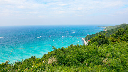 The image shows a beautiful coastal scene with turquoise waters, boats anchored near the shore, and lush green hills in the background.