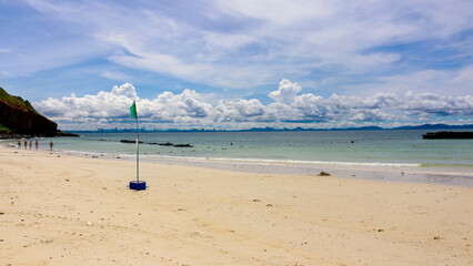 A serene beach scene with a green flag on the sandy shore, gentle waves meeting the coastline, a rocky hill to the left, and distant cityscape under a cloudy sky. koh larn,thailand