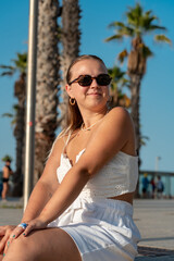 Blond young girl in white outfit and sunglasses traveling in Europe. Palm trees and blue sky on background in Barcelona in summer.