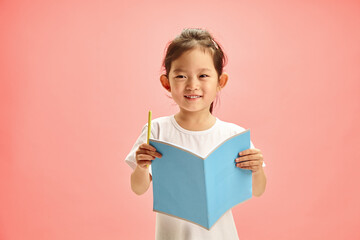 Happy and Pretty Preschool-age Korean Girl with an open Notebook with Cover Facing out and Holding Pen, Smiling Sweetly Standing on a Pink Isolated Background. Concept of Remote Learning.