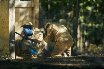 Paintball player in protective mask aims gun, takes cover under fire, paint splashes. Paintball...