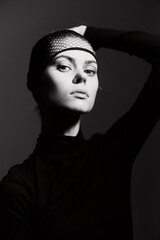 Elegant woman in black turtleneck with fishnet headpiece posing dramatically against a dark background, showcasing confidence and contemporary fashion aesthetics.