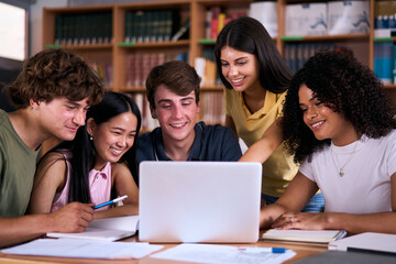 Diverse group of high school young students working together using and looking at laptop in library. Happy Generation Z classmates multiracial friends studying sitting with computer on class project