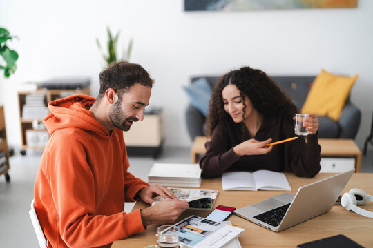 Two young adults collaborating at a desk, surrounded by books, a laptop, and work materials. The casual atmosphere suggests teamwork or studying, with one person pointing to something in a book