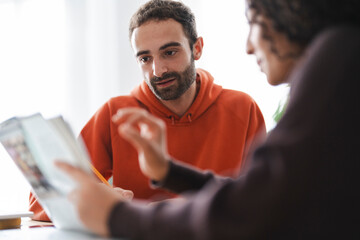 man and woman sit at a table with a laptop and books as they brainstorm ideas. They are having an in-depth conversation in a bright and modern space, symbolizing teamwork and creative collaboration.