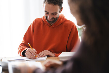 A man in an orange hoodie writes in a notebook at a table, surrounded by books and papers, fully engaged in a focused learning environment.