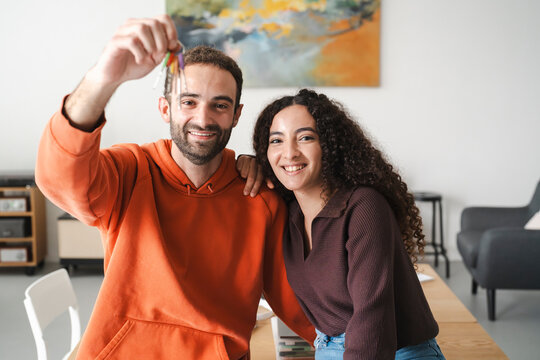 A cheerful young couple sitting together at a table, with the man in an orange hoodie holding up a set of keys, symbolizing a fresh beginning or new home in a brightly lit, modern room.